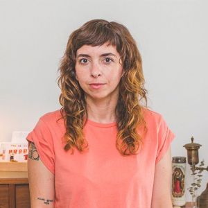 a white nonbinary person with long brown hair sits facing the camera directly in a coral-colored shirt