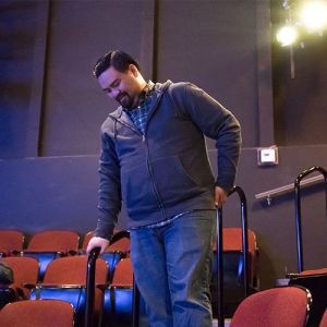 A brown skinned Filipino American person in a grey hoodie on the steps of a theatre looking toward the ground.