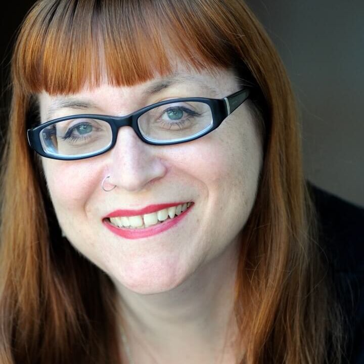 Headshot of Melissa Hillman. A light skinned woman with blue eyes, black oval glasses and wearing red lipstick with a nose ring. She has red hair with bangs. She is smiling with her teeth showing.