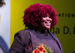 Rolanda D. Bell, a Black woman with red curls, smiles in the spotlight, holding a bouquet of flowers.