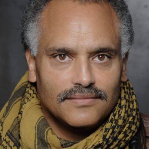 Headshot of Michael Gene Sullivan. A light skinned black man with light brown eyes, short curly white and grey hair and a mustache. He is wearing a yellow and black scarf