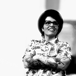 Black and white photo of Dani Spinks. SHe is wearing a patterned button up and glasses. Her arms are crossed and she has tattoos on her arms