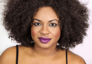 Headshot of Tanika Baptiste. She is a Black woman with shoulder length natural curly brown hair. She is staring directly at the camera and wearing purple lipstick.