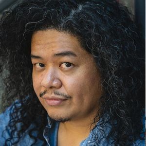 A portrait photo of artist, Tasi Alabastro. He's a Filipino man wearing a blue button-up shirt. He's at a three-quarter angle head turned towards the camera, eyes making direct contact with the lens. His curly hair came down past his jawline, framing his face.