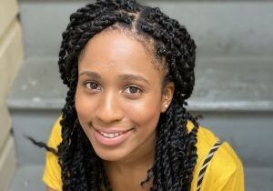 Angel sits smiling on a grey staircase wearing a yellow shirt and black and white striped pants. She is a black woman with waist length dark hair styled in twists.