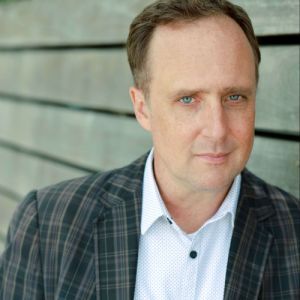 Picture of a white man with a serious expression, wearing a white shirt and Dark Grey Checked Jacket