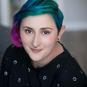 A shoulders-up photo of a white woman in her mid thirties with a slight smile. She is wearing a black t-shirt with a pattern of white o’s on. She has a nose ring, gauged ears and rainbow hair. The background (a grey sofa) is blurred out of focus.